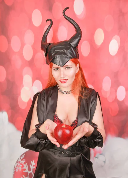 Руда дівчина в сексуальній чорній сукні з диявольськими рогами тримає червоне яблуко з спокусою і спокусою — стокове фото