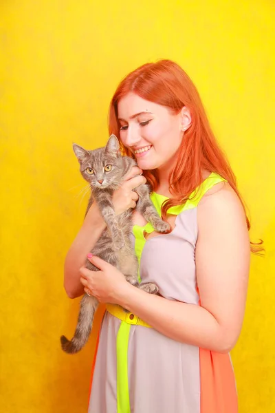 Красивая молодая рыжая девушка в длинном красочном летнем платье с ее прекрасной подругой серый кот на желтом ярком студийном фоне — стоковое фото