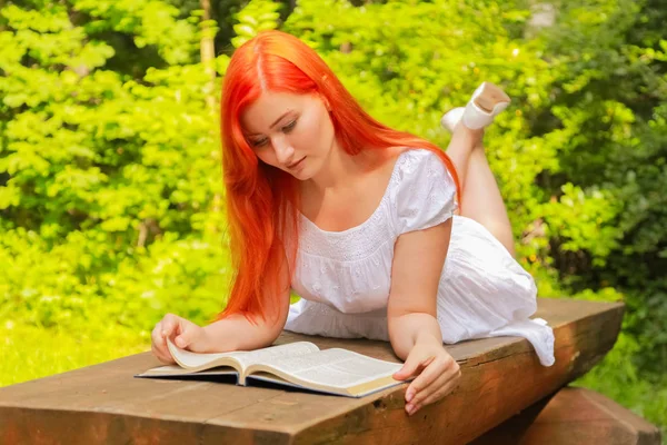 Adolescente com cabelo vermelho em algodão branco vestido de descanso e leitura do livro em um verão parque da cidade quente — Fotografia de Stock