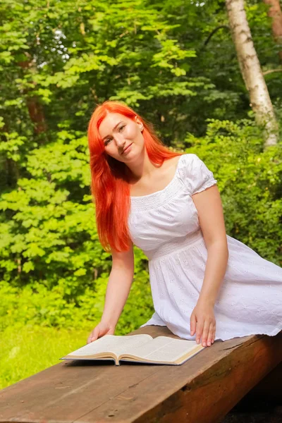 Έφηβο κορίτσι με κόκκινα μαλλιά σε λευκό βαμβακερό φόρεμα ανάπαυσης και διαβάζοντας το βιβλίο σε ένα καλοκαιρινό ζεστό πάρκο της πόλης — Φωτογραφία Αρχείου