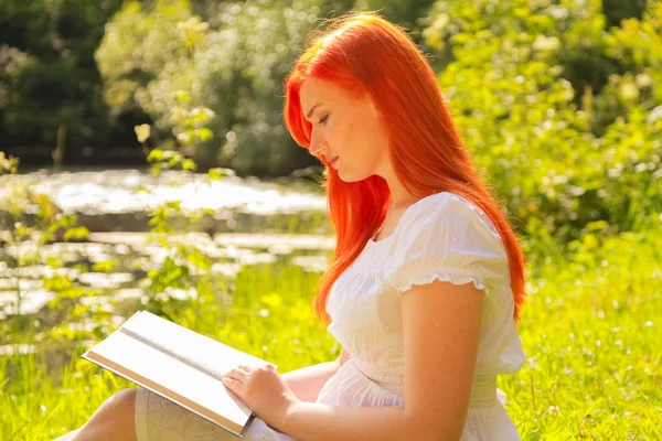 Adolescente com cabelo vermelho em algodão branco vestido de descanso e leitura do livro em um verão parque da cidade quente — Fotografia de Stock