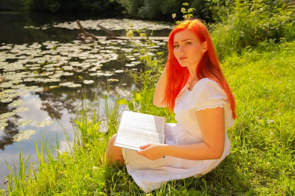 Дівчина-підліток з рудим волоссям у білій бавовняній сукні відпочиває і читає книгу в літньому гарячому міському парку — стокове фото