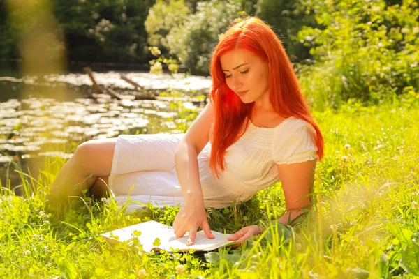 Tizenéves lány vörös hajú, fehér pamut ruhában pihenés és a könyv olvasása egy nyári forró Városliget — Stock Fotó