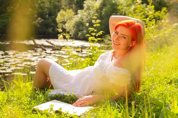Adolescente com cabelo vermelho em algodão branco vestido de descanso e leitura do livro em um verão parque da cidade quente — Fotografia de Stock