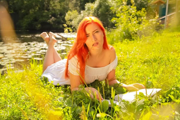 Mladá dívka s červenými vlasy v bílých bavlněných šatech a četbou knihy v letním horkém městském parku — Stock fotografie