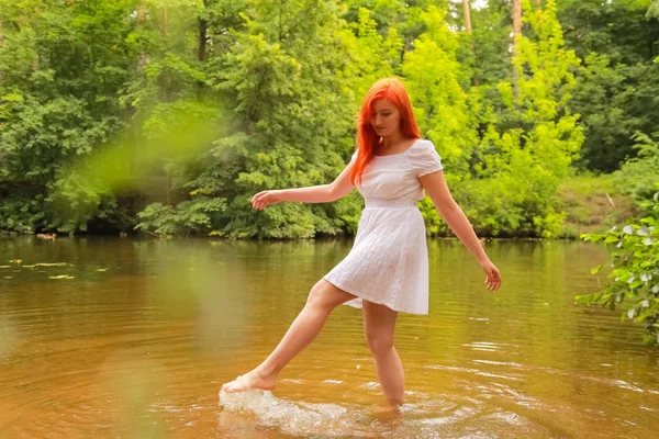 Mulher ruiva feliz em vestido branco em um rio se divertindo e salpicando água no verão. pessoa feliz brinca com água a pé . — Fotografia de Stock