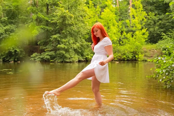 Ευτυχισμένη κοκκινομάλλα γυναίκα με λευκό φόρεμα σε ένα ποτάμι που διασκεδάζει και να πιτσιλίζει το νερό το καλοκαίρι. ευτυχισμένο άτομο παίζει με το νερό με τα πόδια. — Φωτογραφία Αρχείου