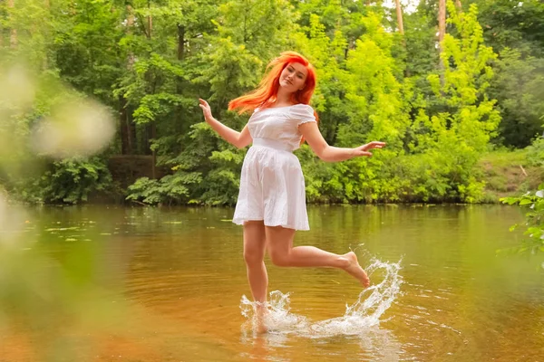 Ευτυχισμένη κοκκινομάλλα γυναίκα με λευκό φόρεμα σε ένα ποτάμι που διασκεδάζει και να πιτσιλίζει το νερό το καλοκαίρι. ευτυχισμένο άτομο παίζει με το νερό με τα πόδια. — Φωτογραφία Αρχείου