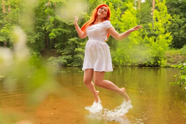Mujer pelirroja feliz en vestido blanco en un río que se divierte y salpica el agua en verano. persona feliz juega con el agua a pie . — Foto de Stock