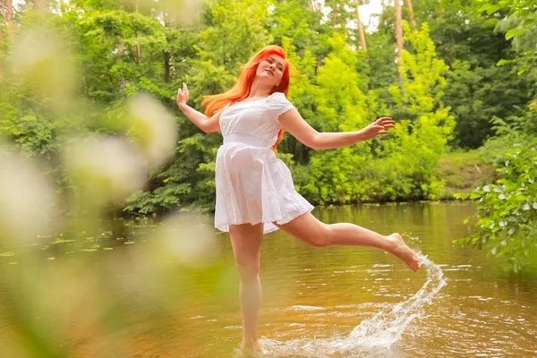 Glückliche rothaarige Frau im weißen Kleid an einem Fluss, die Spaß hat und im Sommer planscht. Glücklicher Mensch spielt zu Fuß mit Wasser. — Stockfoto