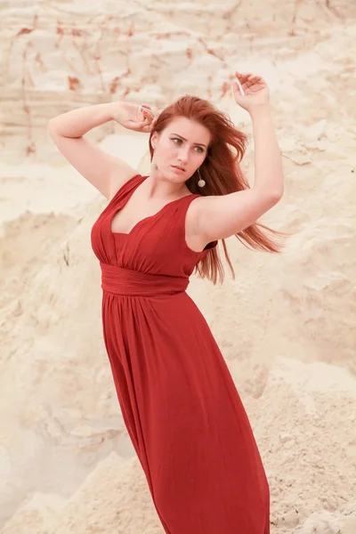 Fiatal szép molett kaukázusi nő, piros, hosszú ruhában pózol a sivatagban táj homokkal. — Stock Fotó