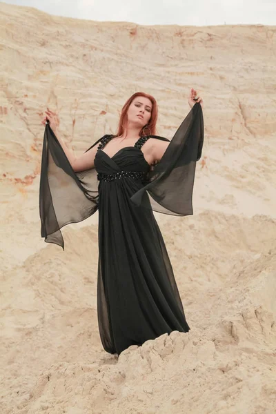 Jeune belle femme caucasienne en robe de mousseline noire posant dans un paysage désertique avec du sable . — Photo