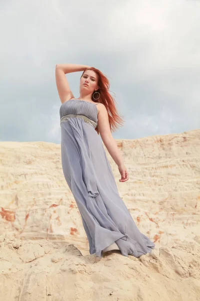 Une fille aux cheveux roux en robe longue flottant dans le vent se tient sur un monticule de sable dans le désert et aime le vent — Photo