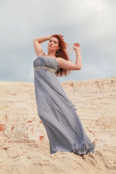 Рыжая девушка в длинном платье, развевающемся на ветру, стоит на насыпи песка в пустыне и любит ветер — стоковое фото
