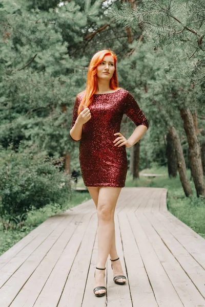 도시 공원 배경에서 걷는 체리 컬러 이브닝 드레스를 입은 빨간 머리의 여성과 여름을 즐기십시오. — 스톡 사진