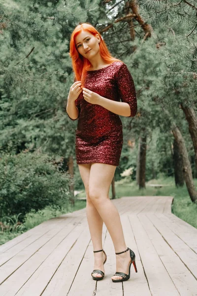 도시 공원 배경에서 걷는 체리 컬러 이브닝 드레스를 입은 빨간 머리의 여성과 여름을 즐기십시오. — 스톡 사진