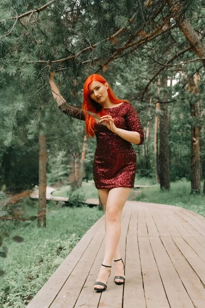 Femme aux cheveux roux en robe de soirée couleur cerise marchant dans le fond du parc de la ville et profiter de l'été — Photo