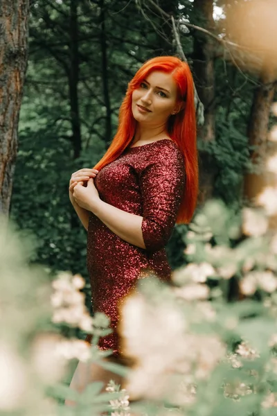 Donna dai capelli rossi in abito da sera di colore ciliegia passeggiando sullo sfondo del parco cittadino e godersi l'estate — Foto Stock