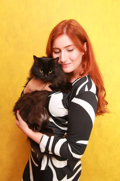 Красивая молодая рыжая девушка в длинном черно-белом платье со своим прекрасным другом кошкой на желтом ярком фоне студии — стоковое фото