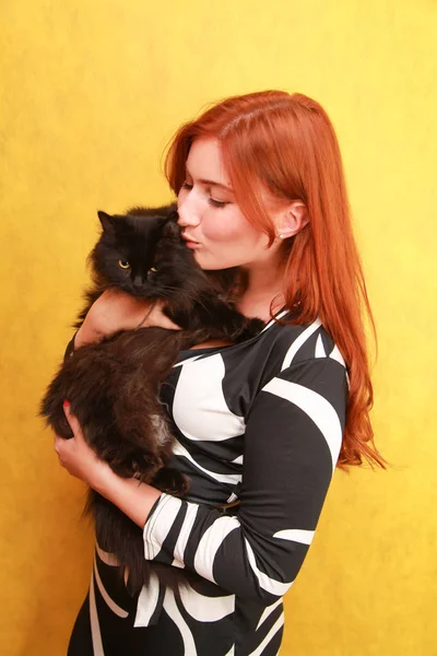 Красивая молодая рыжая девушка в длинном черно-белом платье со своим прекрасным другом кошкой на желтом ярком фоне студии — стоковое фото