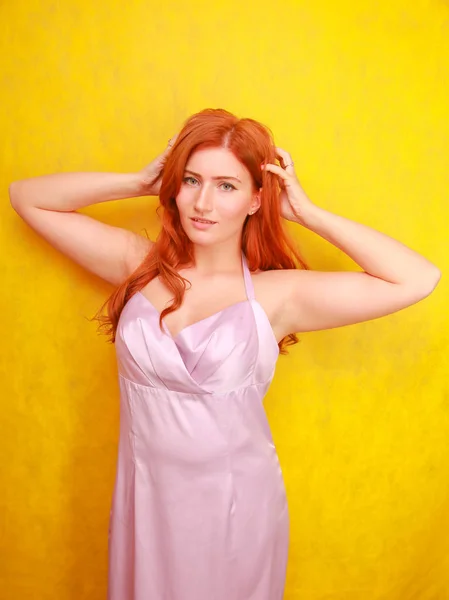 Mooie schoonheid rood haar vrouw in roze zijden jurk op gele achtergrond alleen. speelse plus size roodharige volwassen meisje. — Stockfoto
