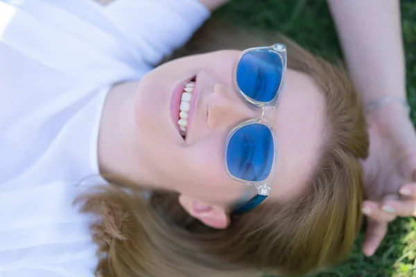 Hübsches kaukasisches Mädchen mit blauer Sonnenbrille liegt auf dem grünen Gras und lacht glücklich — Stockfoto