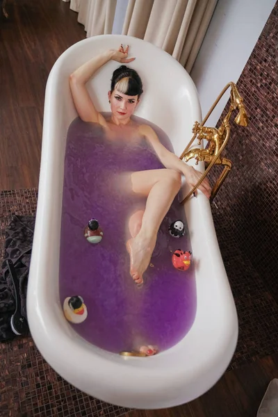 Здоровая готическая девушка с черными волосами в роскошной коричневой ванной с фиолетовой соленой водой — стоковое фото