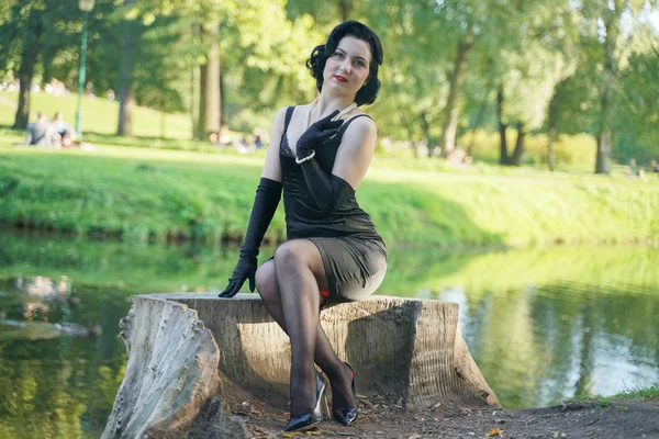 Ritratto esterno della ragazza pinup seduta sul ceppo nel parco cittadino vicino all'acqua — Foto Stock