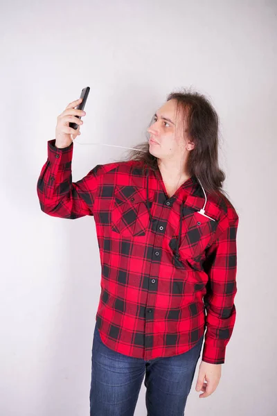 Hombre nerd de pelo largo en una camisa roja a cuadros con un teléfono sobre un fondo blanco en el estudio — Foto de Stock