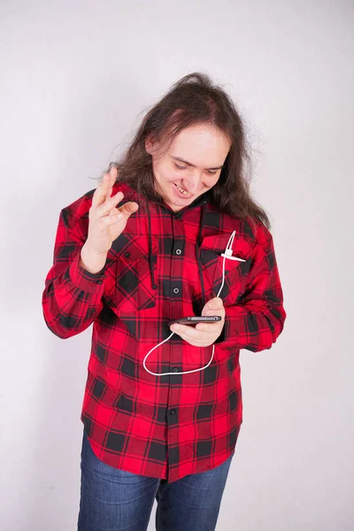 Hombre nerd de pelo largo en una camisa roja a cuadros con un teléfono sobre un fondo blanco en el estudio — Foto de Stock