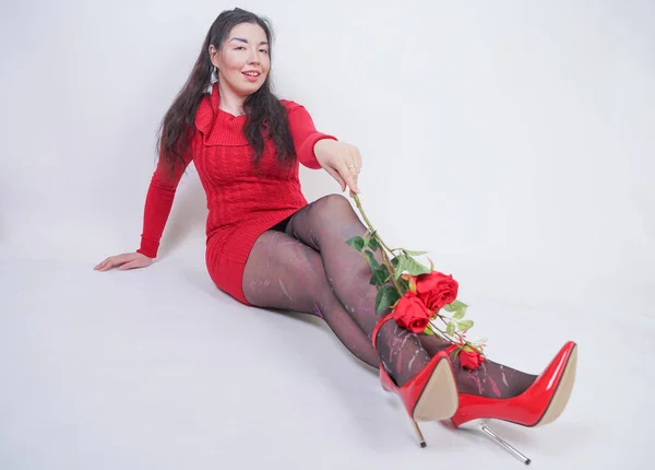 Grazioso mix corse plus size ragazza in elegante abito rosso midi seduto su sfondo bianco studio con rosa rossa — Foto Stock