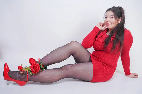 Grazioso mix corse plus size ragazza in elegante abito rosso midi seduto su sfondo bianco studio con rosa rossa — Foto Stock