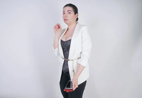 Kouzelný mixový závod plus velikostní žena v bílém podnikatelovém kabátku, který je nešťastný a smutný s chytrou telefonem v ruce na bílém pozadí ve studiu — Stock fotografie