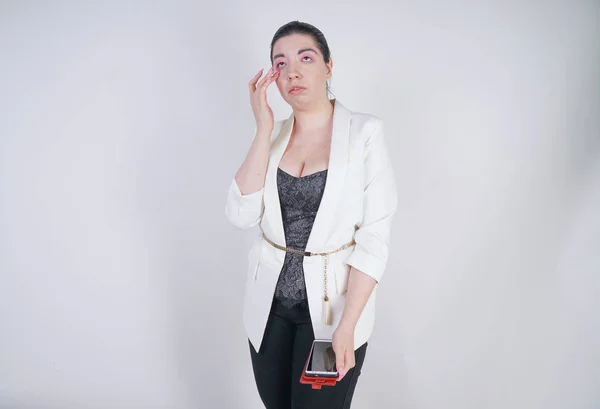 Γοητευτικό αγώνα Mix Plus μέγεθος γυναίκα σε ένα λευκό επιχειρηματικό μπουφάν στέκεται δυστυχισμένη και λυπημένος με έξυπνο τηλέφωνο στο χέρι σε λευκό φόντο στο στούντιο — Φωτογραφία Αρχείου