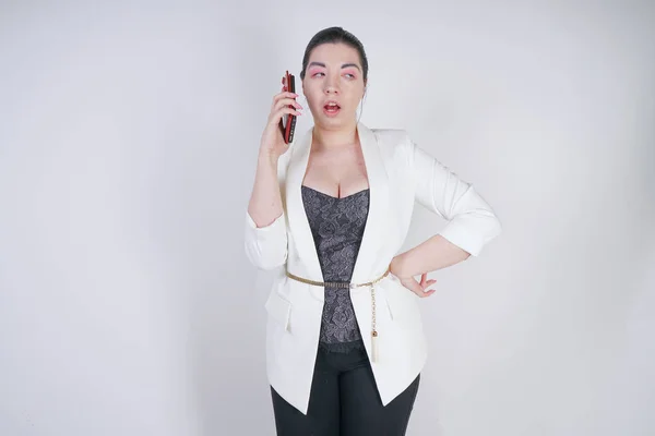 スタジオで白い背景に手にスマートフォンを持って立っている白いビジネスジャケットで魅力的なミックスレースプラスサイズの女性 — ストック写真
