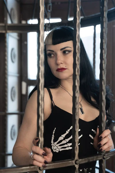 Sexy gothic girl in tight black dress behind the big cage posing as slavery victim alone — Stock Photo, Image