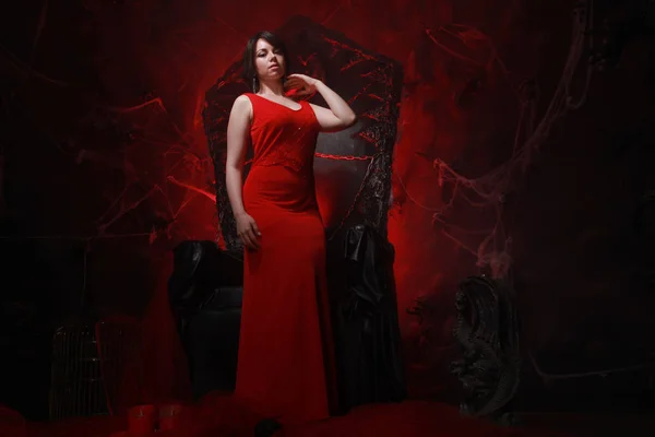 Schöne einsame Mädchen in langen roten Kleid auf dem Halloween-Thron bereit für gruselige Party — Stockfoto