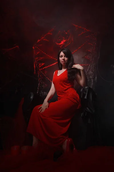 Красивая одинокая девушка в длинном красном платье на Хэллоуинском троне готова к страшной вечеринке — стоковое фото