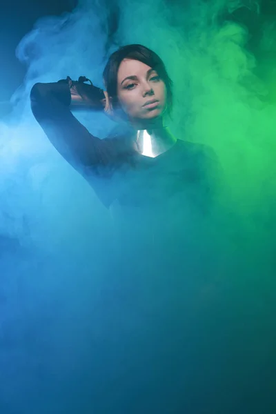 Modelo de moda futurista con ropa negra y plateada y de pie en el colorido humo azul y verde —  Fotos de Stock