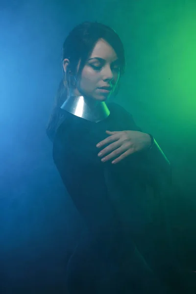 Футуристична модель моди в чорно-срібному одязі і стоїть в барвистому синьо-зеленому димі — стокове фото