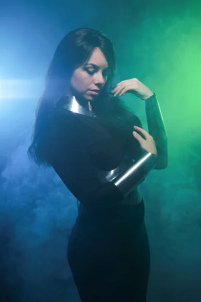 Futuristic fashion model wearing black and silver clothes and standing in the colorful blue and green smoke — Stock Photo, Image