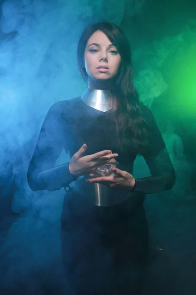 Futuristisk mode modell klädd i svart och silver kläder och stående i färgglada blå och grön rök — Stockfoto