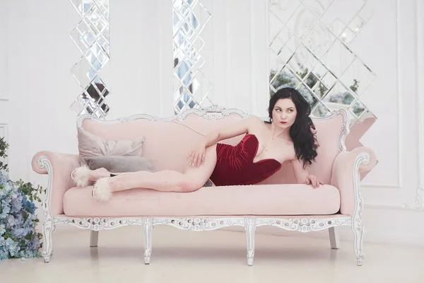 Bastante feliz hembra en terciopelo rojo body posando en el interior con rosa retro sofá — Foto de Stock