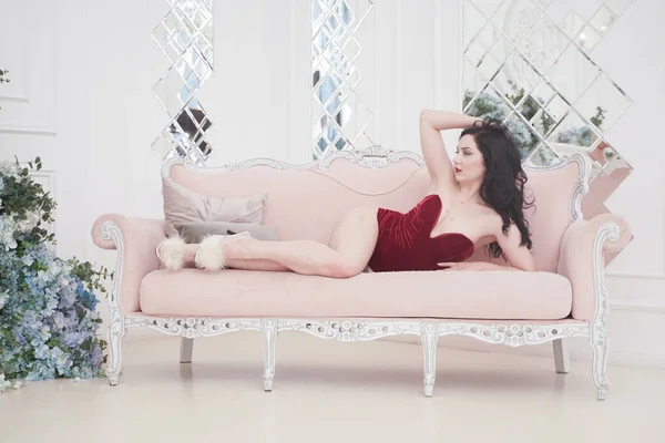 Bastante feliz hembra en terciopelo rojo body posando en el interior con rosa retro sofá — Foto de Stock