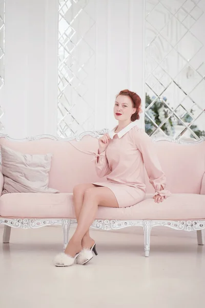 Young beautiful caucasian woman posing in pink retro dress indoors alone. slim adult girl in vintage outfit with old sofa on white background. — Stock Photo, Image