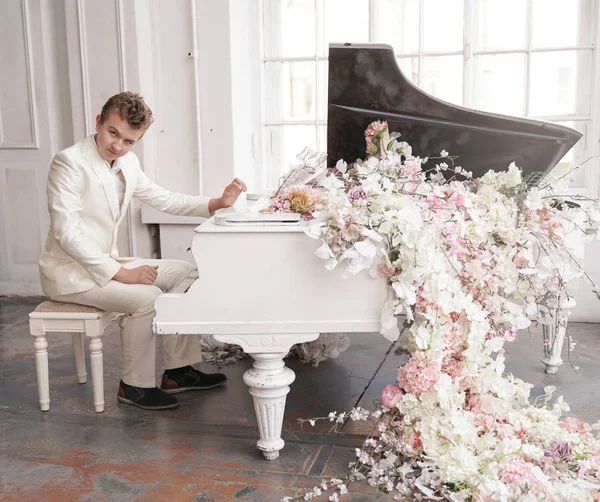 Ung man Tonåring i en vit affärs dräkt med ett stort vitt piano i blommor. kaukasiska manliga spela musik på semester ensam. — Stockfoto