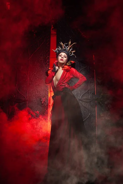 Kırmızı duman ile büyük bir kapının arka planda uzun lüks bir elbise ağaç kökleri şeklinde boynuzları ile güzel moda cadı kadın. Cadılar Bayramı konsepti — Stok fotoğraf