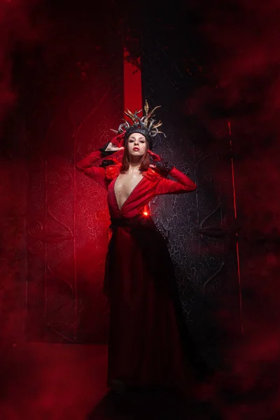 Hermosa mujer bruja de la moda con cuernos en forma de raíces de árbol en un vestido largo y lujoso en el fondo de una enorme puerta con humo rojo. Concepto Halloween —  Fotos de Stock