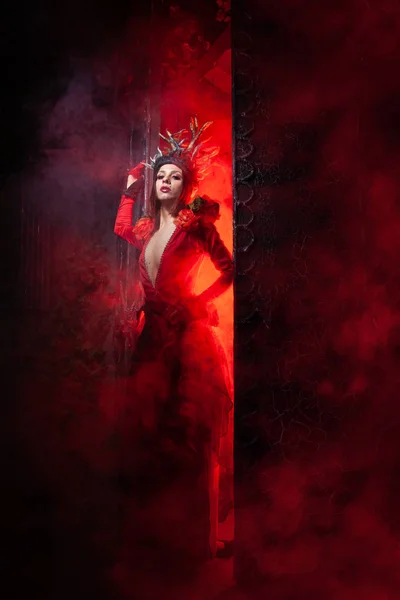 Hermosa mujer bruja de la moda con cuernos en forma de raíces de árbol en un vestido largo y lujoso en el fondo de una enorme puerta con humo rojo. Concepto Halloween —  Fotos de Stock