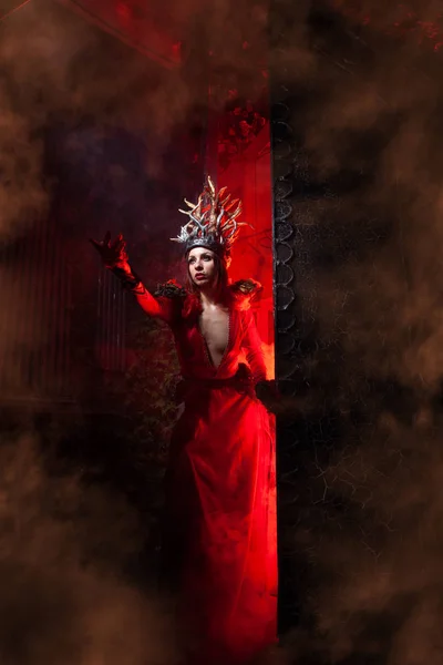 Piękna dziewczyna czarownica mody z rogami w postaci korzeni drzew w długiej luksusowej sukni na tle ogromnej bramy z czerwonym dymem. Koncepcja Halloween — Zdjęcie stockowe